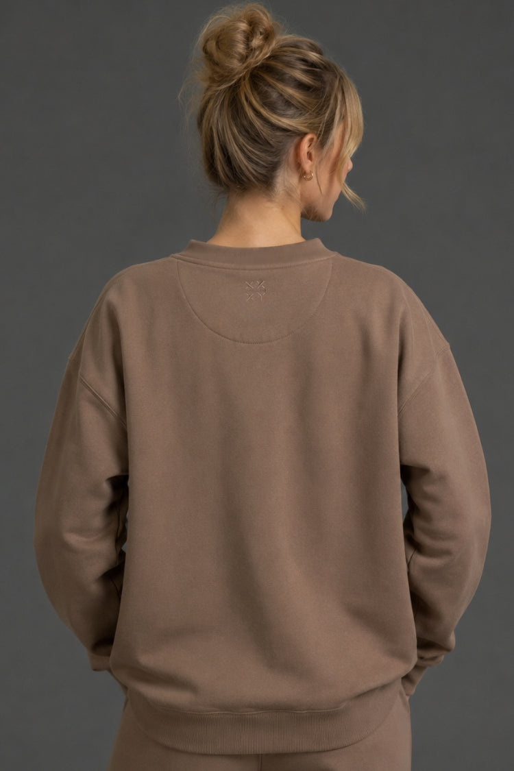A woman viewed from behind wearing a taupe brown crewneck sweatshirt and matching sweatpants against a dark gray studio background. || Mocha