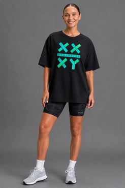 Female model wearing the black XX-XY Athletics graphic T-shirt with black bike shorts on a gray studio background. || Black