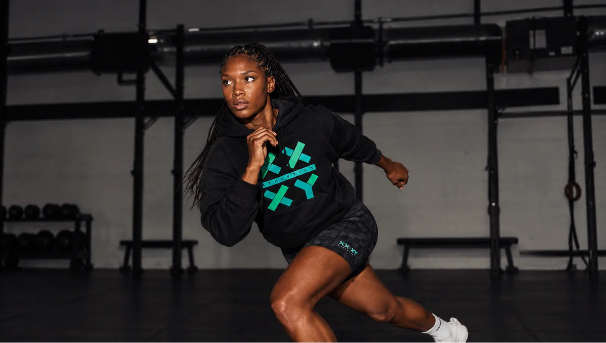 A woman in a black athletic hoodie with a teal geometric logo and black shorts poses dynamically in a gym setting.