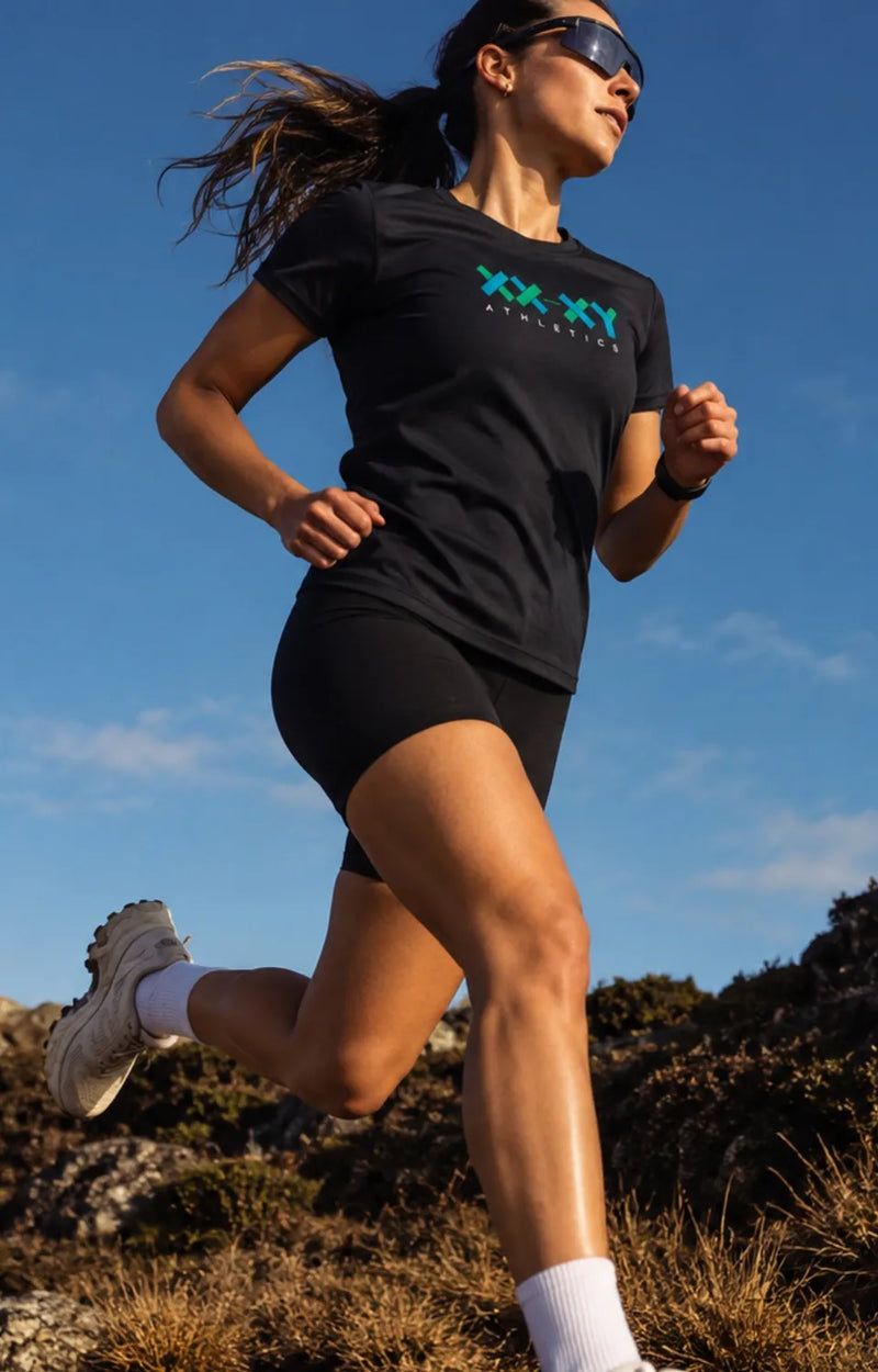 A woman running outdoors in a black athletic t-shirt with a teal and white 'XX-XX Athletics' logo, paired with black shorts, white socks, and running shoes under a clear blue sky.