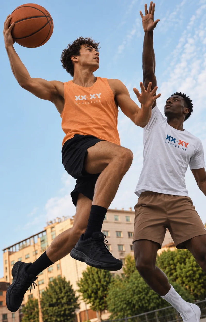 Two men playing basketball outdoors under a blue sky. The man in the foreground is wearing an orange sleeveless tank top with 'XX-XY' branding, black shorts, and black sneakers, while holding a basketball.