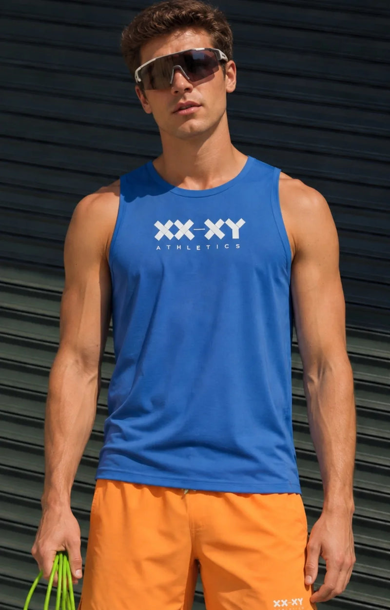 A man wearing a bright blue sleeveless athletic tank top with a white 'XX-XY ATHLETICS' logo on the chest, paired with orange athletic shorts, standing in front of a dark corrugated metal wall.