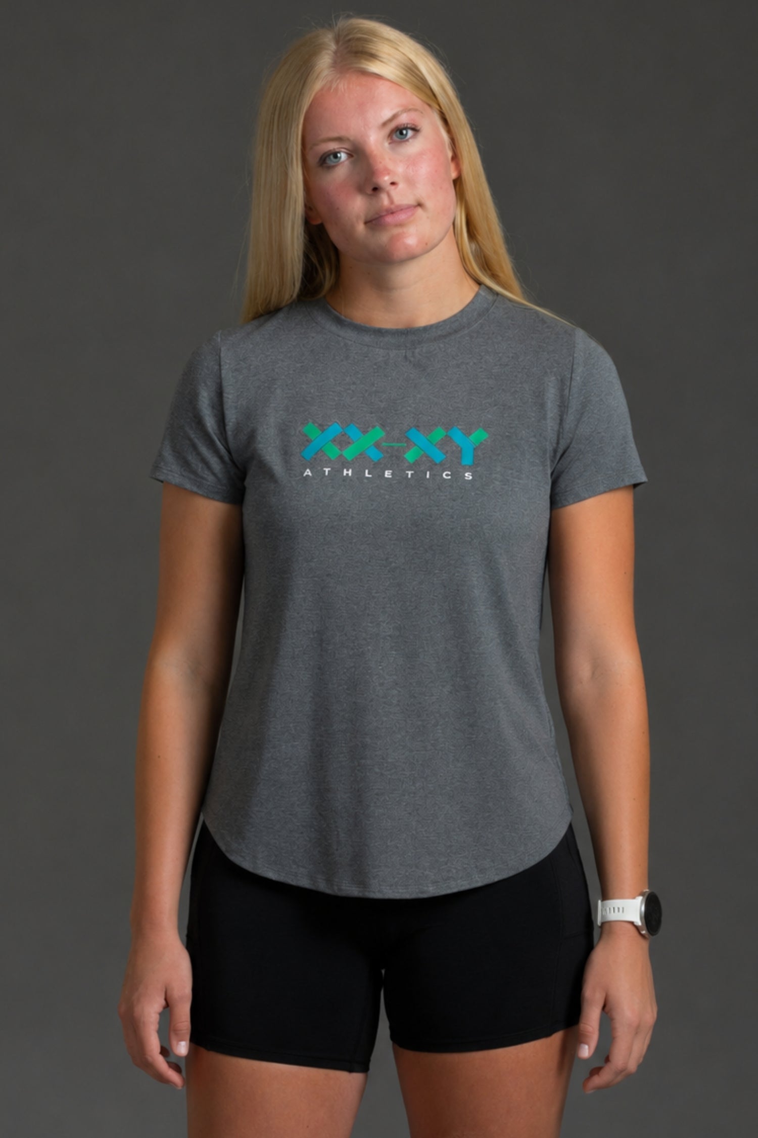 A woman modeling a heather gray athletic t-shirt with a teal and white 'XX XY ATHLETICS' logo on the chest, paired with black athletic shorts, against a dark gray studio background.