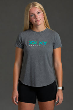 A woman modeling a heather gray athletic t-shirt with a teal and white 'XX XY ATHLETICS' logo on the chest, paired with black athletic shorts, against a dark gray studio background. || Dark Heather Gray