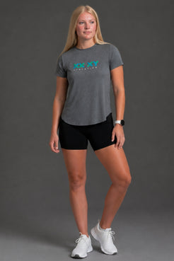 A woman modeling a heather gray athletic t-shirt with a teal and white 'RUN KY RUN' graphic on the chest, paired with black athletic shorts and white sneakers, against a solid gray studio background. || Dark Heather Gray