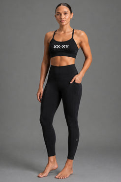 A woman modeling a black XX-XY activewear set, featuring a sports bra with a white logo and matching high-waisted leggings, against a solid gray studio background. || Black Heather
