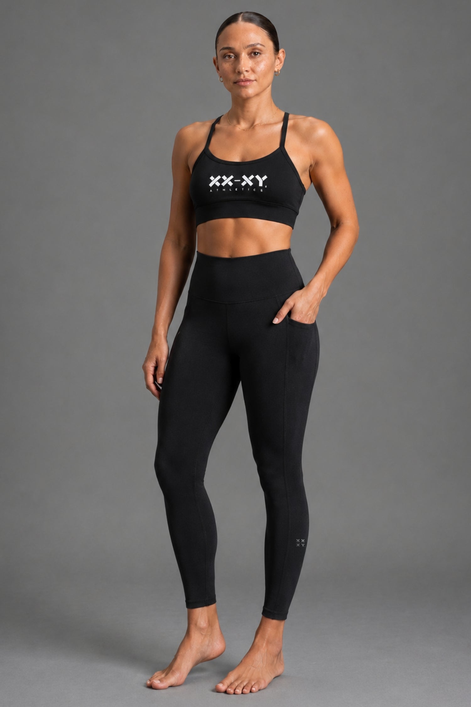 A woman modeling a black XX-XY activewear set, featuring a sports bra with a white logo and matching high-waisted leggings, against a solid gray studio background. || Black Heather