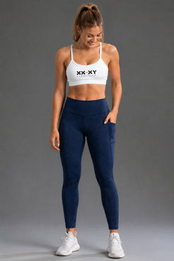 A woman modeling a white sports bra with a black 'XX-XY' logo and navy blue high-waisted leggings with side pockets, paired with white sneakers, against a solid gray studio background. || Navy