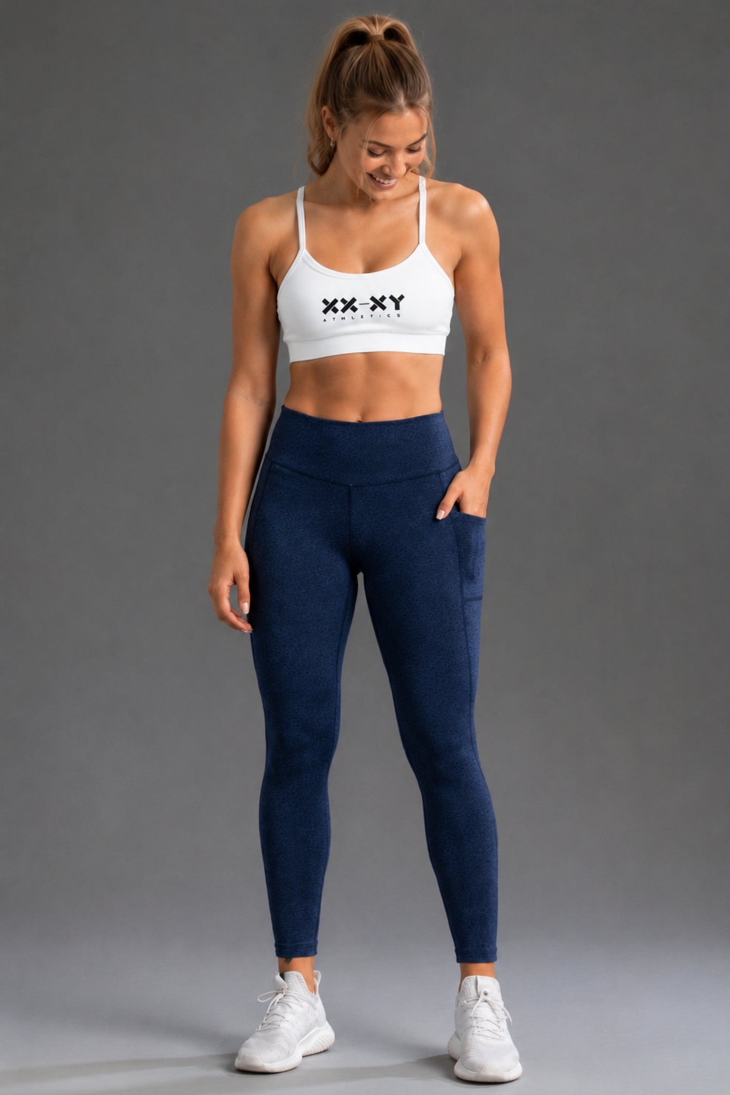A woman modeling a white sports bra with a black 'XX-XY' logo and navy blue high-waisted leggings with side pockets, paired with white sneakers, against a solid gray studio background. || Navy