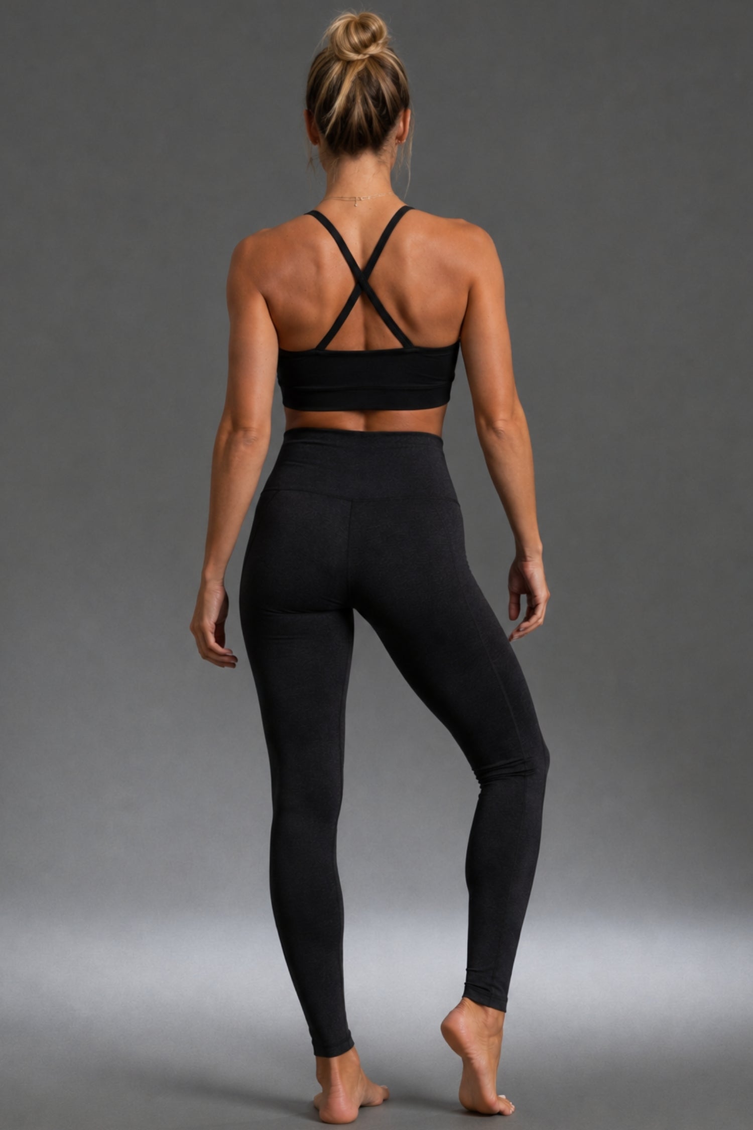 A woman is shown from behind wearing a black sports bra with crisscross back straps and black high-waisted leggings, standing against a neutral gray studio background.