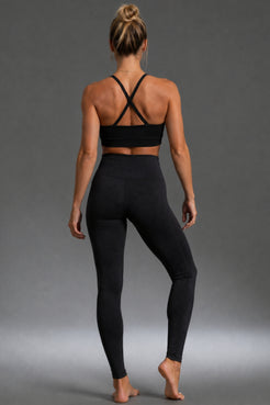 A woman is shown from behind wearing a black sports bra with crisscross back straps and black high-waisted leggings, standing against a neutral gray studio background. || Black Heather