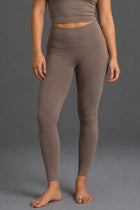 A model wearing high-waisted, ankle-length leggings in a solid taupe-brown color, shown from the waist down against a neutral gray studio background. || Mocha