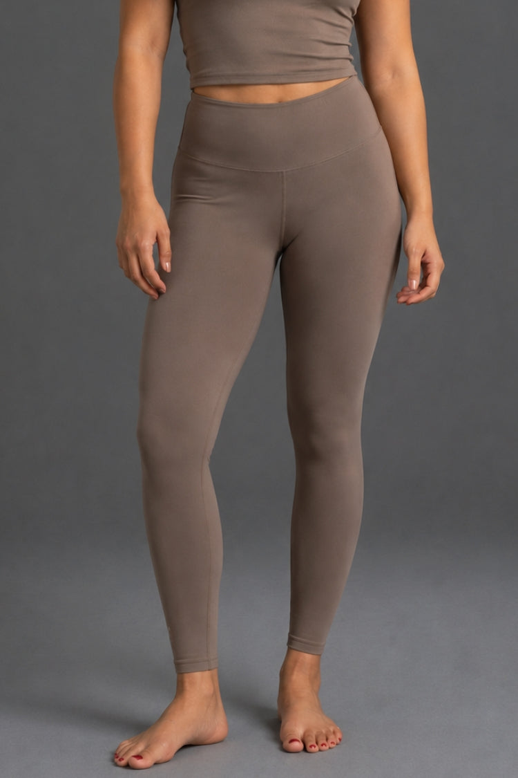 A model wearing high-waisted, ankle-length leggings in a solid taupe-brown color, shown from the waist down against a neutral gray studio background.