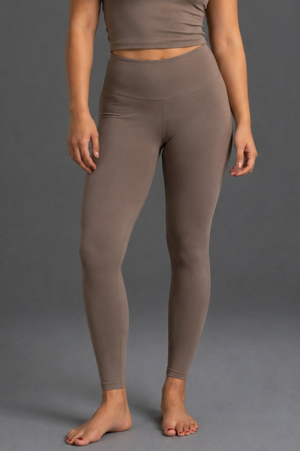 A model wearing high-waisted, ankle-length leggings in a solid taupe-brown color, shown from the waist down against a neutral gray studio background. || Mocha