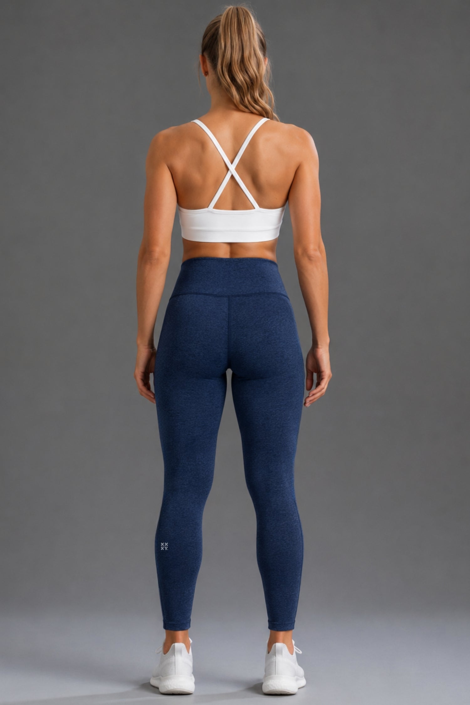 A woman shown from the back wearing navy blue high-waisted leggings and a white sports bra, standing against a solid gray studio background.