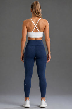 A woman shown from the back wearing navy blue high-waisted leggings and a white sports bra, standing against a solid gray studio background. || Navy