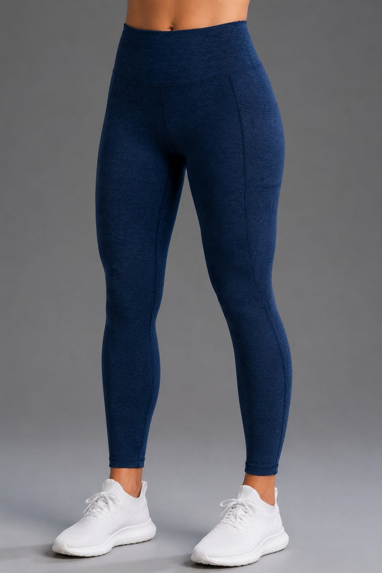 A person wearing high-waisted navy blue leggings and white athletic sneakers, shown from the waist down against a plain gray studio background. || Navy
