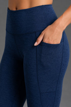 Close-up of a person wearing heathered navy blue high-waisted leggings with a side pocket, showing the hand in the pocket against a neutral gray background. || Navy