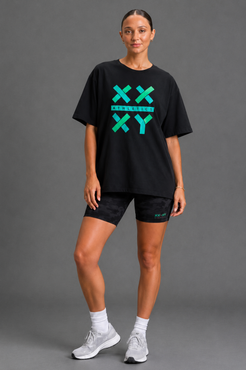 A woman modeling a black athletic t-shirt with a bright green 'XX XY' graphic on the front, paired with black shorts, white socks, and white sneakers, against a solid gray studio background. || black