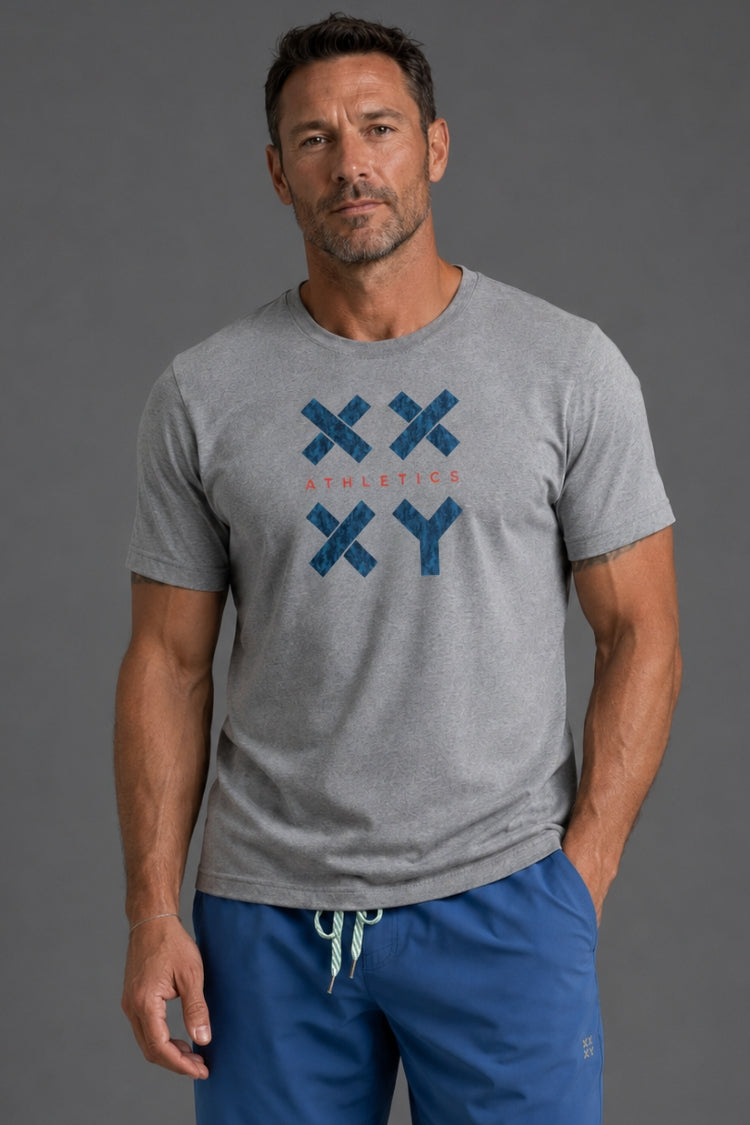 A man wearing a heather gray athletic t-shirt with a blue 'XXY' logo and 'ATHLETICS' text on the chest, paired with blue drawstring sweatpants, against a solid gray studio background.