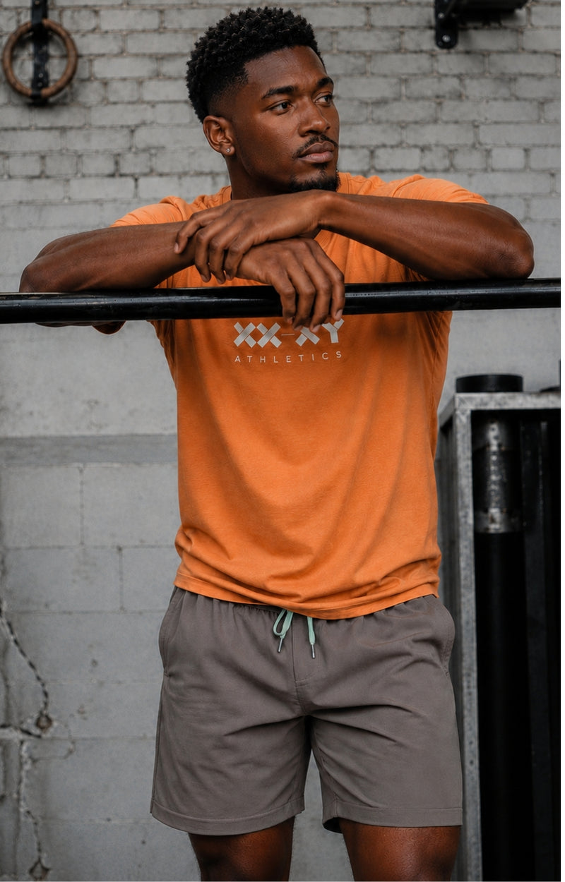 A man wearing an orange athletic t-shirt with 'XXI XY ATHLETICS' printed on the chest and gray drawstring shorts, leaning on a black bar in front of a gray brick wall.