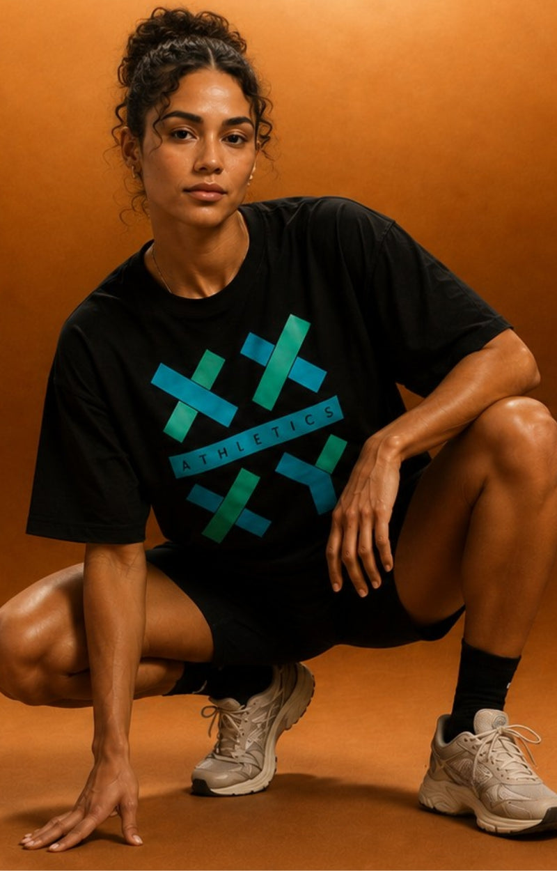 A woman crouching in a studio setting wearing a black athletic t-shirt with a teal and blue geometric cross graphic and the word 'ATHLETICS' across the chest, paired with black shorts and gray running shoes against a warm brown background.