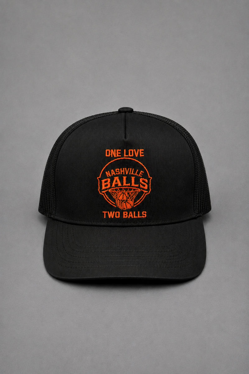 A black trucker-style baseball cap with orange embroidered text and a basketball graphic on the front, photographed against a neutral gray background. || Black