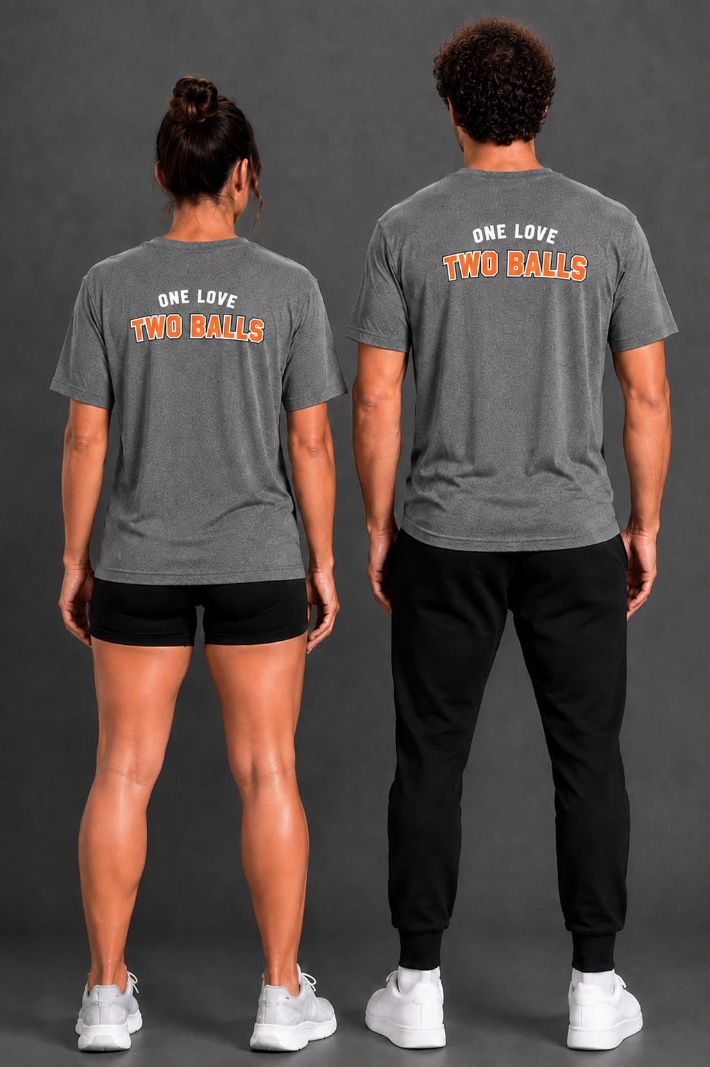 Two models shown from the back wearing matching dark gray t-shirts with the text 'ONE LOVE TWO BALLS' printed in navy blue and orange. The model on the left wears black athletic shorts and white sneakers, while the model on the right wears black jogger pants and white sneakers, set against a dark gray studio background. || Dark Heather Gray