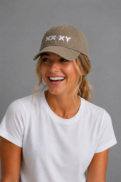 A woman smiling while wearing a khaki baseball cap with a white embroidered logo that reads 'XXXY AESTHETICS' and a white t-shirt, set against a solid gray studio background. || Mocha