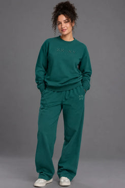 Woman wearing a pine green XX-XY Athletics crewneck sweatshirt and matching wide-leg sweatpants against a gray studio background. || Pine