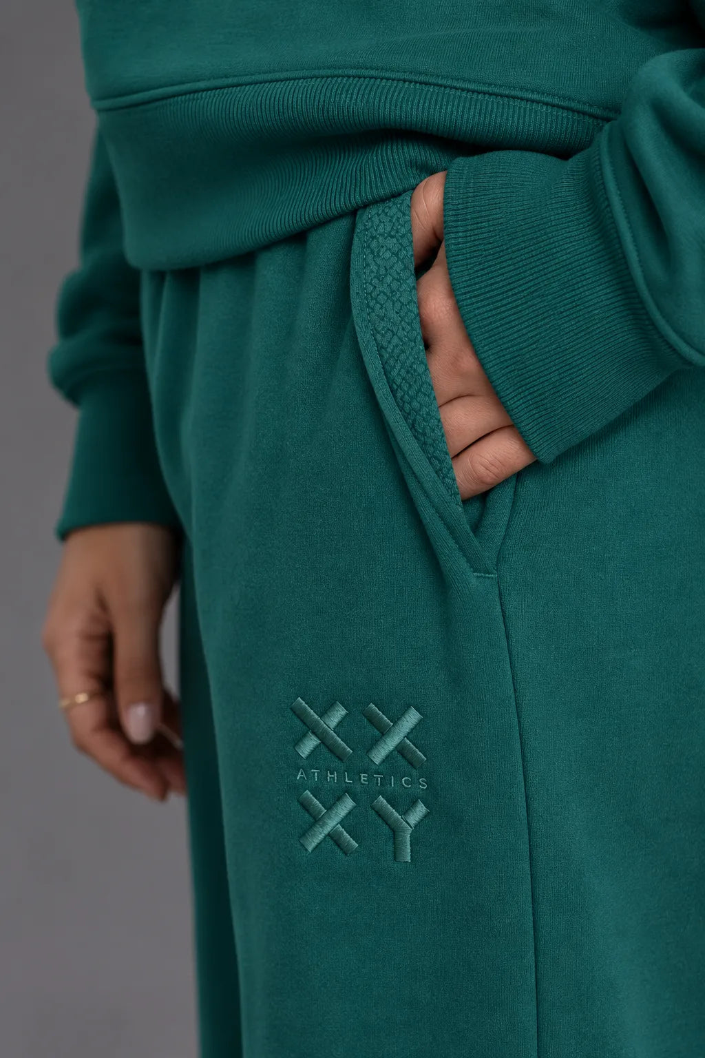 Close-up of pine green XX-XY Athletics sweatpants featuring a tonal embroidered logo and side pocket detail. || Pine