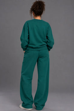 A person viewed from behind wearing a matching dark teal-green sweatshirt and wide-leg sweatpants set, standing against a plain gray studio background. || Pine