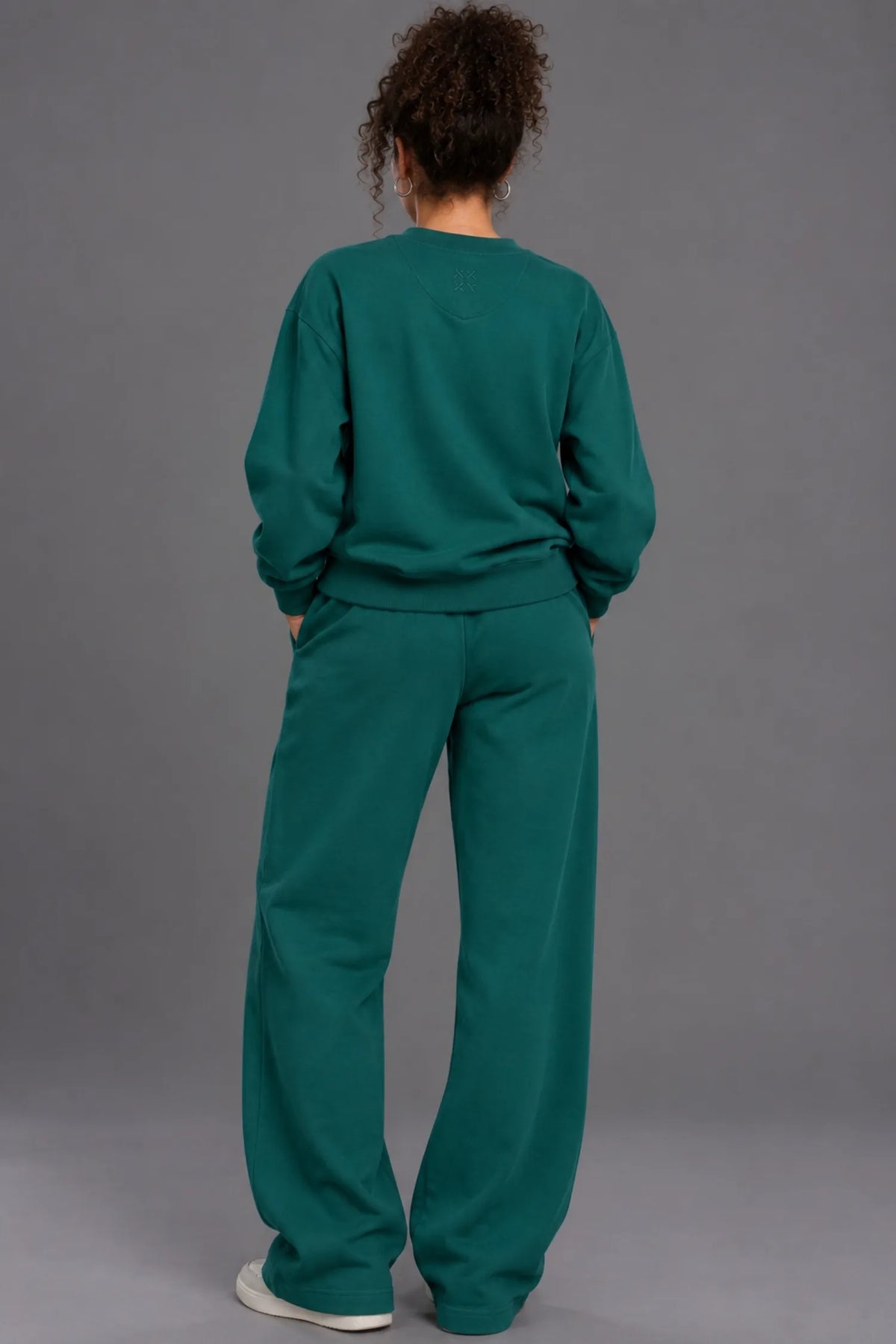 A person viewed from behind wearing a matching dark teal-green sweatshirt and wide-leg sweatpants set, standing against a plain gray studio background. || Pine