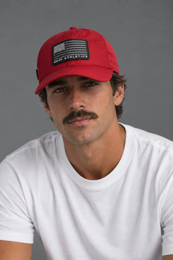 A man wearing a red baseball cap with a black American flag patch and 'NAVY ATHLETICS' text, paired with a white t-shirt against a solid gray background. || Red