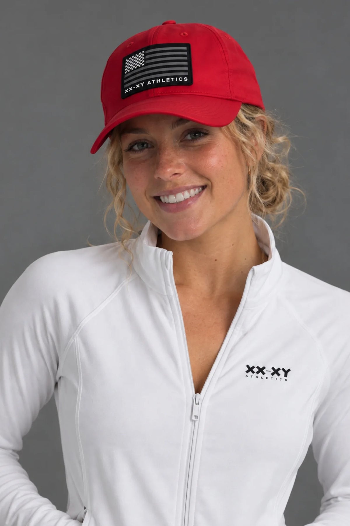 A woman wearing a red baseball cap with a black American flag patch and 'XX-XY ATHLETICS' text, paired with a white zip-up athletic jacket featuring a black 'XX-XY' logo on the chest, against a solid gray background. || Red