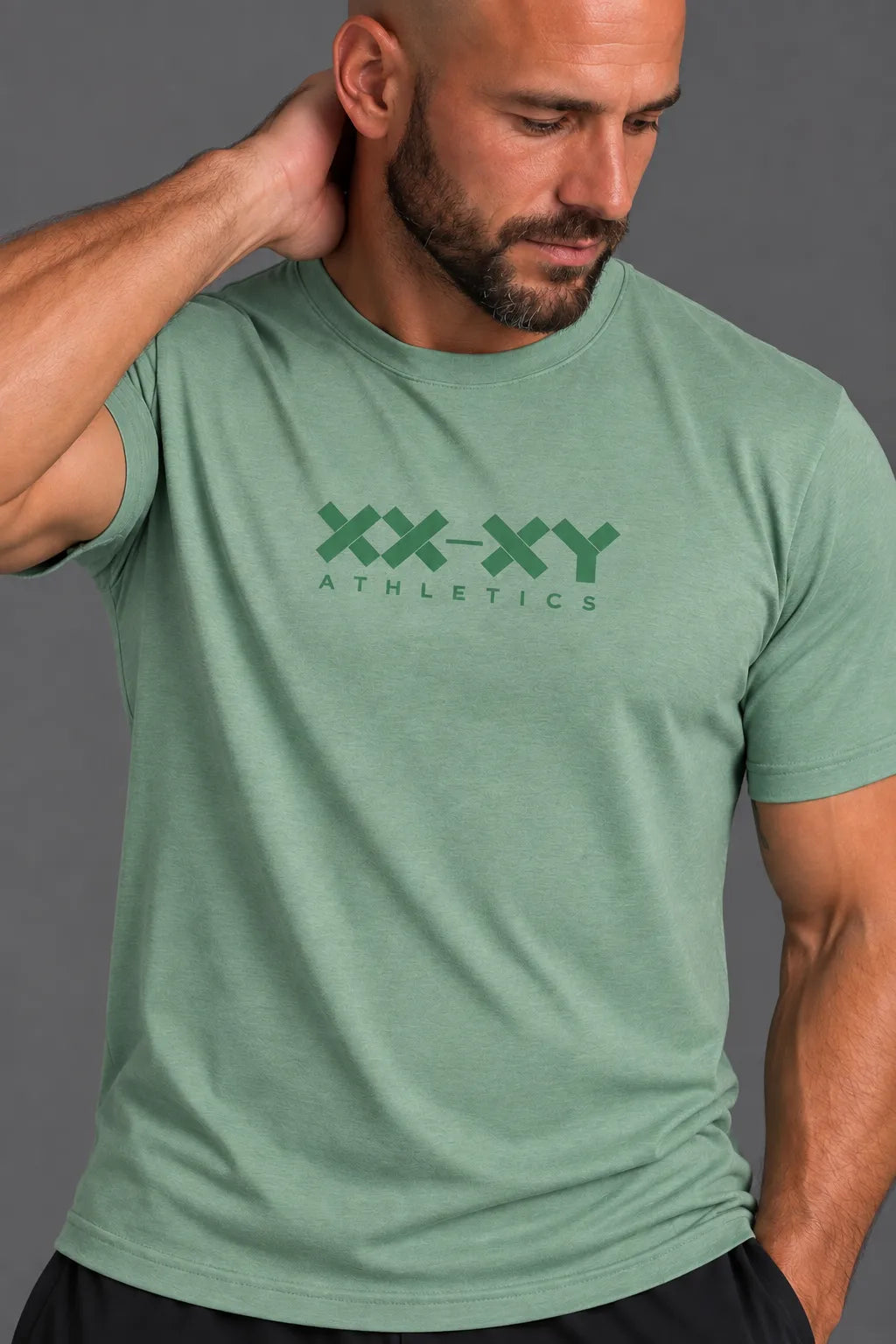 A close-up of a man wearing a heathered sage green t-shirt with a dark green 'XX-XY ATHLETICS' logo printed on the chest, set against a solid gray background.