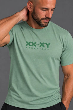 A close-up of a man wearing a heathered sage green t-shirt with a dark green 'XX-XY ATHLETICS' logo printed on the chest, set against a solid gray background. || Sage Heather