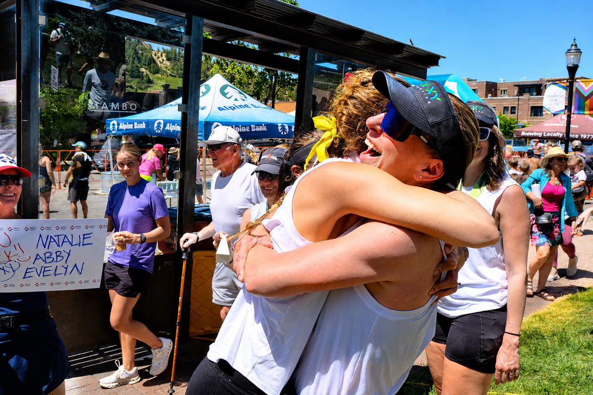 Two women embrace in a crowded outdoor public space on a sunny day. One wears a white tank top and black cap, and the other has curly hair and a yellow top.