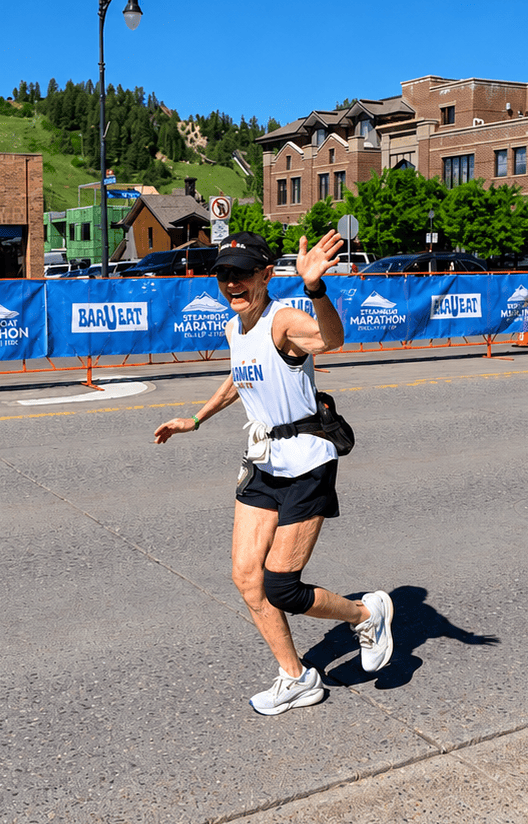 A man running during a marathon on a paved street, wearing a white tank top with blue lettering, black shorts, white running shoes, and a black cap. He is waving to the camera.