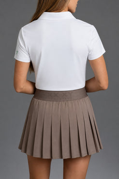 Back view of a woman wearing a white short-sleeve polo shirt and a pleated taupe-brown skirt against a solid gray studio background. || Mocha