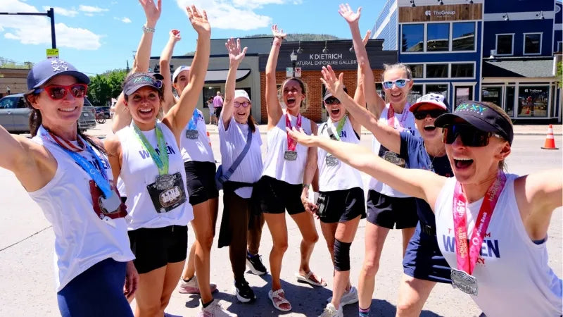 A group of women celebrating outdoors with raised arms, wearing matching white tank tops, black athletic shorts, and race bibs, with one in the foreground reading "WOMEN" on her top.