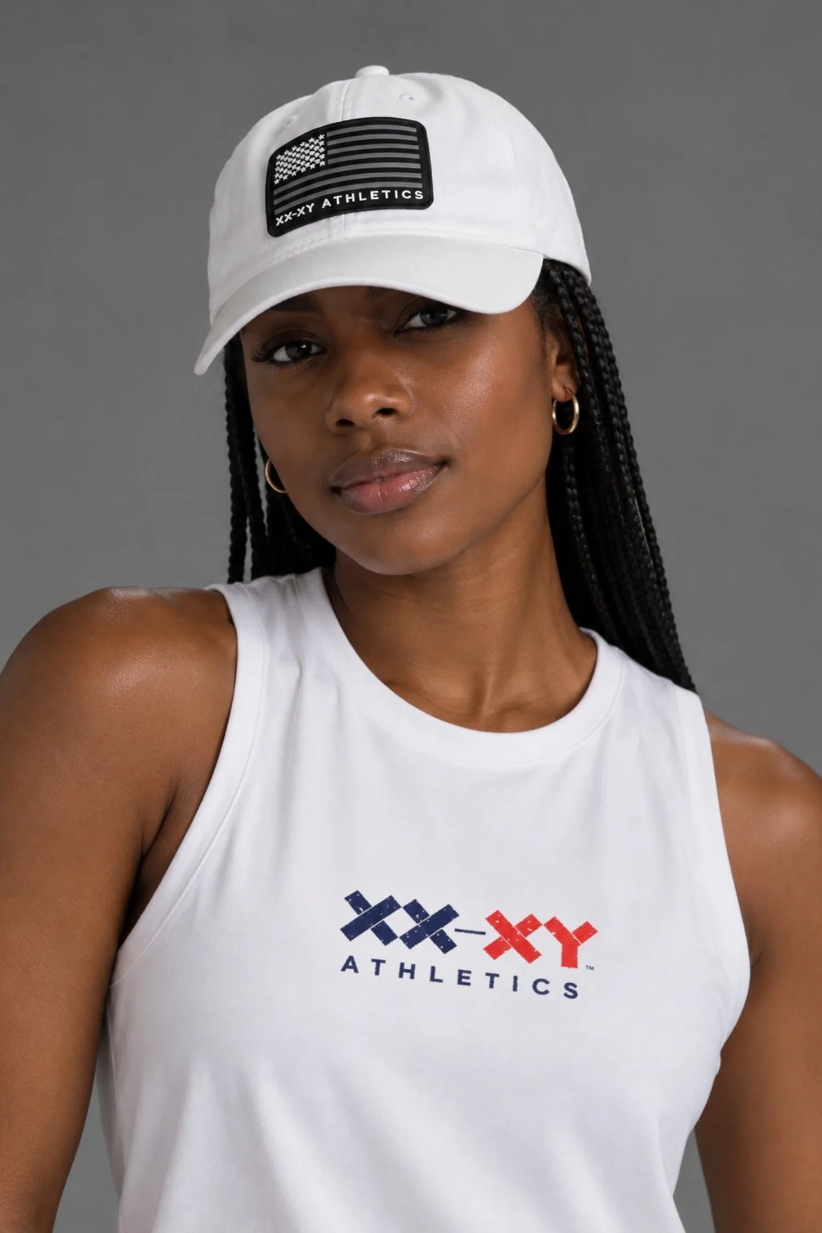 A woman wearing a white baseball cap with a black American flag patch and 'XX-XXY ATHLETICS' text, paired with a white sleeveless tank top featuring a red and blue 'XX-XXY ATHLETICS' logo on the chest, against a solid gray studio background.