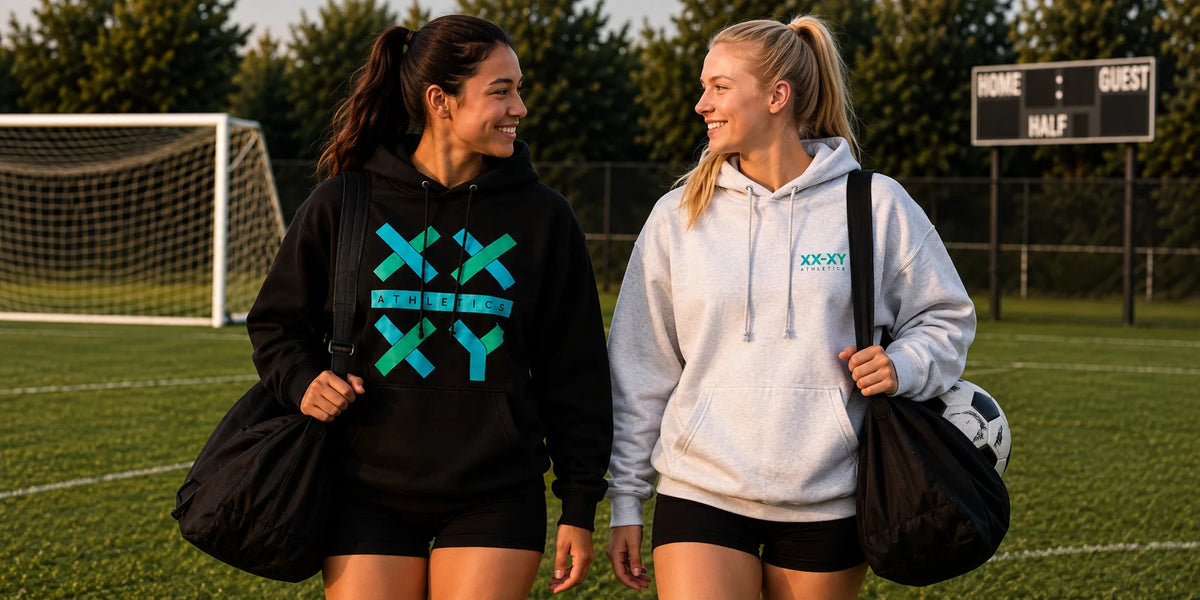 Two female athletes walking and smiling on a sports field, each carrying a black duffel bag. The athlete on the left wears a black hoodie with a teal graphic, and the athlete on the right wears a light gray hoodie with a teal logo.