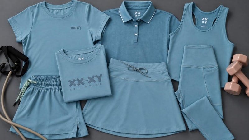 A coordinated athletic apparel set in dusty blue, laid out on a dark gray background. The set includes a short-sleeve T-shirt, a collared polo shirt, a sleeveless tank top, shorts, a skirt, and leggings, with a jump rope and dumbbells as styling props.